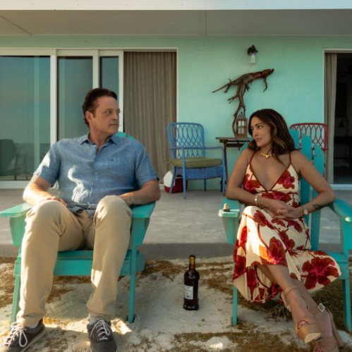 Fotografía en plano medio de dos personas sentadas en sillas de playa de color turquesa frente a una casa color azul pastel. A la izquierda, un hombre con camisa azul de manga corta y pantalones claros (Vince Vaughn) mira hacia la derecha. A la derecha, una mujer con un vestido floreado en tonos rojos y blancos (Natalie Martinez) lo observa mientras conversa. Entre ambas sillas hay una botella en la arena.
