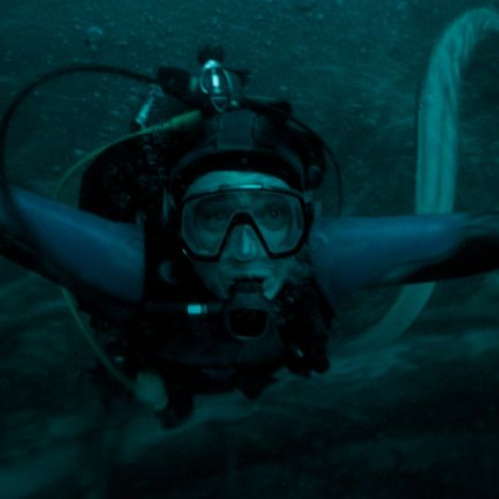 Fotografía en formato horizontal ambientada bajo el agua en tonos azul oscuro y verdoso. En el centro, el actor Austin Abrams aparece caracterizado como buzo, vistiendo un traje de neopreno con visor y boquilla de oxígeno mientras extiende los brazos nadando en medio de aguas profundas.