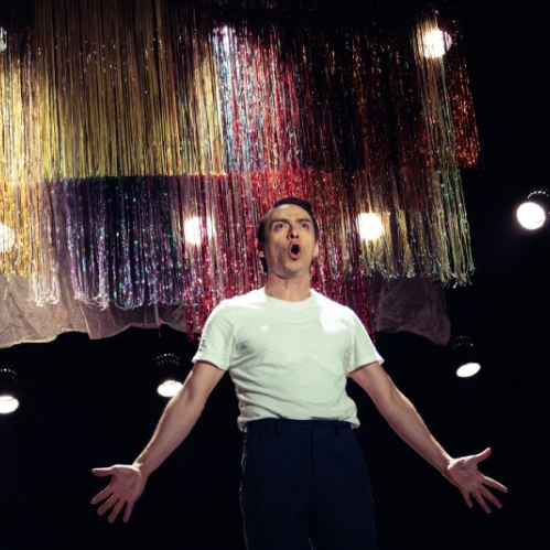Persona sobre un escenario iluminado por reflectores, con brazos extendidos y decoraciones metálicas suspendidas en el fondo.