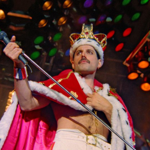 Fotografía en formato horizontal de una presentación en vivo de Queen. Al centro, el vocalista Freddie Mercury aparece de perfil usando una corona dorada con detalles rojos y armiño sobre la cabeza, luciendo una capa roja de felpa con bordes blancos sobre el torso descubierto y pantalones blancos. Sostiene el pedestal de su micrófono inclinado hacia abajo mientras las luces del escenario en tonos amarillos, verdes y rojos iluminan el fondo.