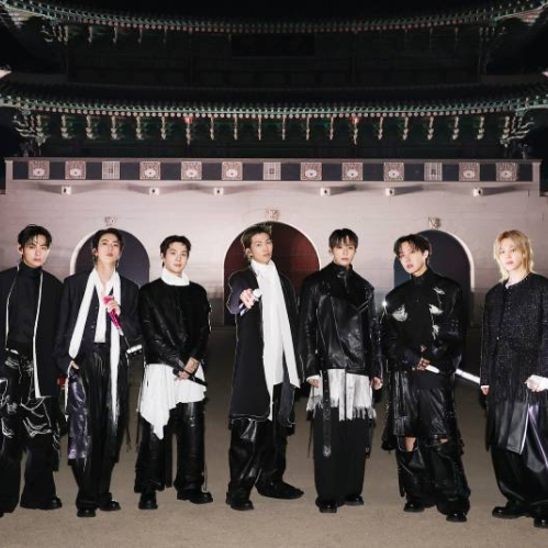 Siete integrantes posan frente a la puerta de Gwanghwamun iluminada de noche, vestidos con atuendos en blanco y negro y sosteniendo micrófonos.