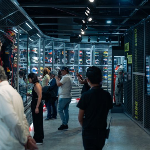 Visitantes recorriendo una sala de exhibición con cascos, trajes y piezas históricas del automovilismo en vitrinas iluminadas.