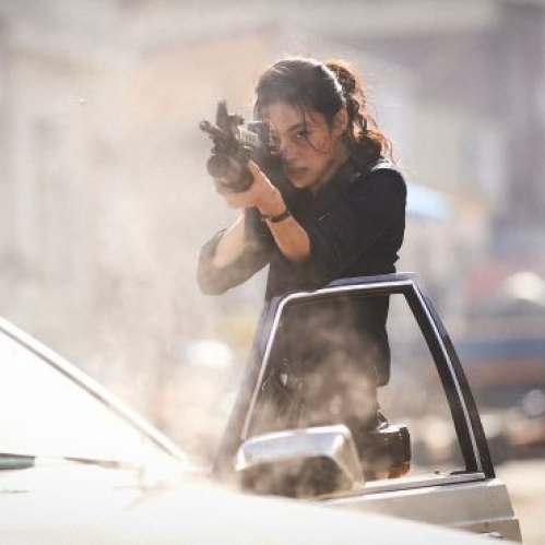 Fotografía fija de la película "Hope: El primer impacto". En una calle luminosa y polvorienta, una mujer apunta con un rifle de asalto desde el costado de una patrulla de policía con la torreta roja encendida. La mujer, con el cabello recogido en una coleta y rastros de suciedad o polvo en el rostro, sujeta el arma con firmeza mientras casquillos de bala vuelan en el aire alrededor de ella en medio de una atmósfera de tensión y acción.