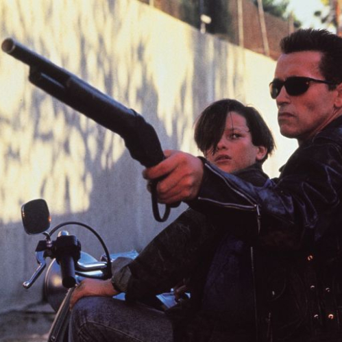 Fotografía en formato horizontal de una escena en exterior durante el día. A la derecha, el actor Arnold Schwarzenegger caracterizado como el T-800 viste una chamarra de cuero negra y gafas de sol oscuras mientras sostiene una escopeta recortada hacia el frente con la mano derecha. Detrás de él, sobre la motocicleta, se observa al actor Edward Furlong en el papel de John Connor, mirando de perfil hacia la escopeta. Al fondo se distingue una barda de concreto clara y palmeras bajo la luz del sol.