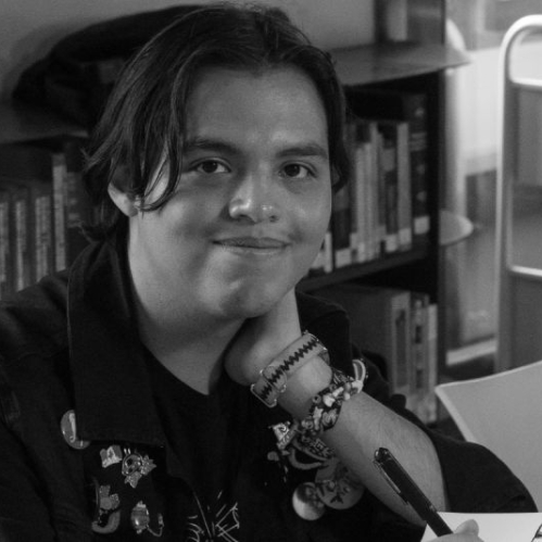 Fotografía en blanco y negro del artista y animador poblano Aarón Cruz sonriendo a la cámara mientras sostiene un bolígrafo sobre su libreta de bocetos. Viste una chamarra con pines y accesorios en la muñeca, sentado en una mesa con libros al fondo.