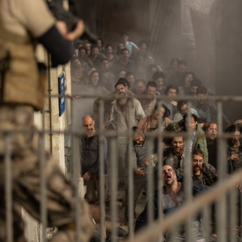 Fotograma de la película Apocalipsis Z: Nuevo Mundo. En el lado izquierdo, en primer plano de perfil y fuera de foco, se observa a un soldado armado sosteniendo un rifle de asalto. A la derecha, al fondo de un pasillo o rampa custodiada por una reja metálica, una multitud enfurecida de infectados se abalanza hacia la cámara con expresiones de desesperación y agresividad.