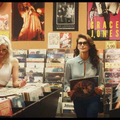 Tres jóvenes con vestimenta de época de los años 80 revisando discos de vinilo dentro de una tienda de música, con pósteres de artistas como Grace Jones, Def Leppard y Prince en la pared de fondo.