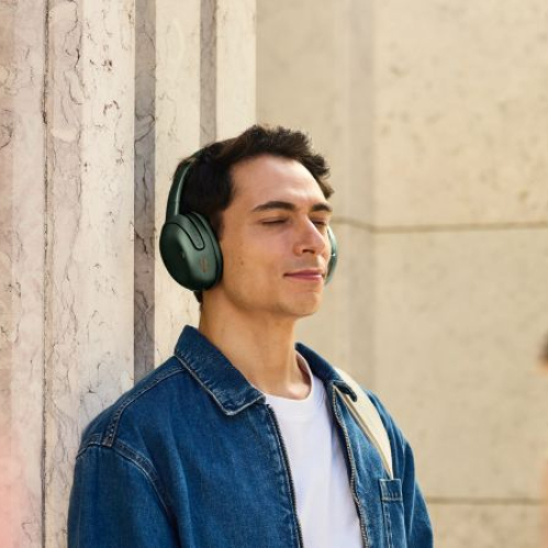 Persona con audífonos verdes de diadema disfrutando audio en un entorno urbano mientras permanece con los ojos cerrados.