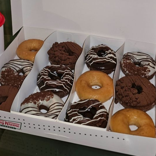 Caja abierta con una docena de donas surtidas colocada junto a un vaso de la marca sobre una superficie de vidrio.