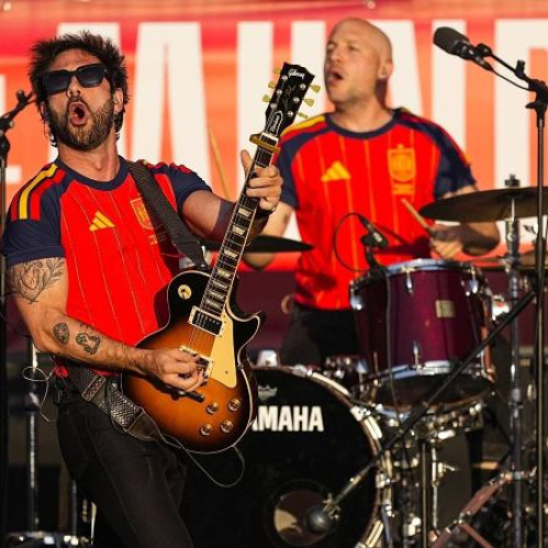 Integrantes de la banda Viva Suecia tocando la guitarra eléctrica y la batería sobre un escenario al aire libre, vistiendo camisetas rojas de la Selección Española de fútbol.