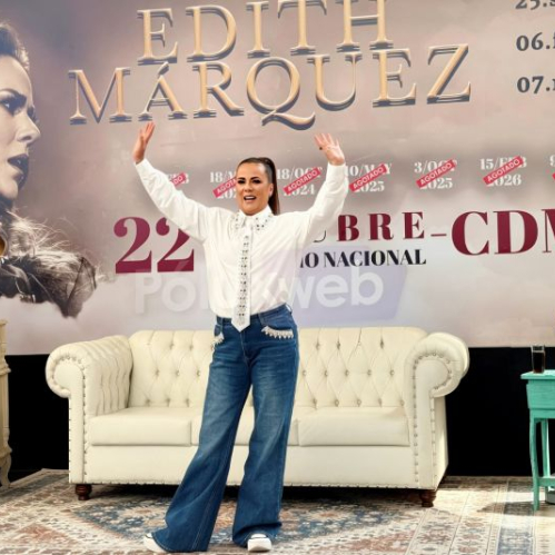 Edith Márquez vestida con camisa blanca decorada y jeans azulosos, posando sonriente con los brazos abiertos frente a la escenografía de su gira en la rueda de prensa.