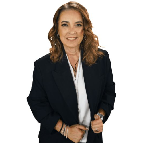 Fotografía de estudio de la periodista y conductora Aurora Valle posando sobre un fondo negro uniforme. Viste un traje formal compuesto por un saco negro holgado sobre una blusa blanca y accesorios metálicos como pulseras en ambas muñecas y cadenas finas al cuello. La presentadora tiene el cabello castaño peinado con ondas suaves sobre los hombros y sostiene la solapa de su saco mientras sonríe levemente a la cámara.