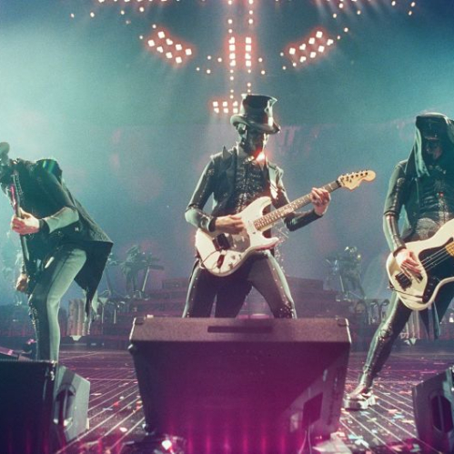 Fotografía en vivo del grupo de metal Ghost durante su presentación en concierto sobre el escenario del Palacio de los Deportes. Tres músicos pertenecientes a los Nameless Ghouls aparecen alineados tocando sus instrumentos bajo una iluminación escénica azul y verde con luces de fondo. Los músicos visten trajes oscuros de época con chaquetas estilizadas, sombreros de copa y máscaras metálicas detalladas. A la izquierda, un guitarrista inclina su instrumento; al centro, otro guitarrista sostiene una guitarra eléctrica blanca modelo Fender Stratocaster; a la derecha, un bajista con postura abierta toca un bajo eléctrico blanco Fender Precision Bass. En el suelo del escenario se observan monitores de retorno de audio negros y confeti esparcido.