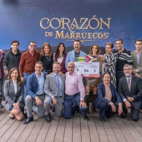Grupo de personas posando frente a un fondo promocional con el título de una producción televisiva, sosteniendo una claqueta durante un evento de inicio de grabaciones.