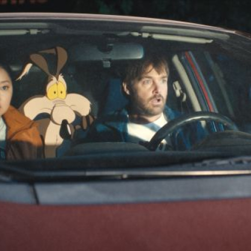 Fotografía de plano medio dentro de un vehículo durante la noche. Al volante se encuentra el actor Will Forte con expresión de sorpresa, mientras a su lado la actriz Lana Condor observa hacia el frente. Entre ambos asientos sobresale la figura animada en dos dimensiones de Wile E. Coyote con gesto preocupado.