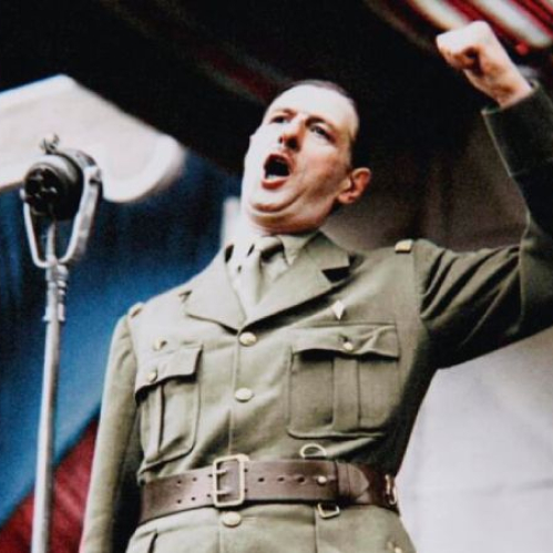Fotografía histórica restaurada a color que muestra en plano medio al general francés Charles de Gaulle luciendo su uniforme militar color caqui y gorro militar. Se encuentra parado frente a dos micrófonos de radio clásicos y una gran estructura decorativa al fondo, alzando con vehemencia el puño derecho mientras exclama un discurso con la boca abierta durante un acto público.