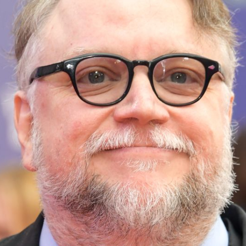 Fotografía de retrato en plano medio del cineasta Guillermo del Toro sonriendo hacia la cámara. Viste una chaqueta oscura, camisa gris claro y lleva sus característicos lentes de armazón negro con barba y cabello canoso. El fondo presenta un tono azul violeta fuera de foco.