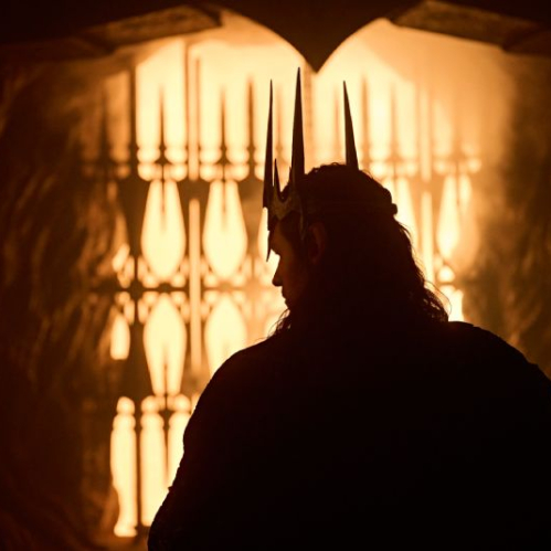 Fotografía cinematográfica a contraluz con tonos cálidos y dorados donde se observa la silueta oscura de espaldas del personaje Sauron, luciendo una corona de espinas o picos metálicos sobre su cabeza y una capa oscura. Al fondo, a través de un arco o portal con rejas de hierro talladas, se aprecia una luz brillante e intensa que evoca fuego y forjas.