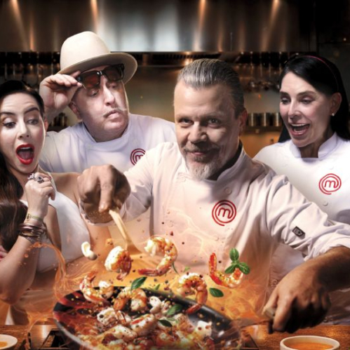 Fotografía publicitaria en formato horizontal para la gran final de MasterChef 24/7. Al centro destaca el chef Poncho Cadena salteando camarones en una sartén con llamas de fuego. A la izquierda, la conductora Claudia Lizaldi expresa sorpresa y a su lado el chef Adrián Herrera observa con sombrero y lentes oscuros. A la derecha, la chef Zahie Téllez sonríe con las manos abiertas. En el fondo se aprecia una cocina profesional con los logotipos oficiales de MasterChef.