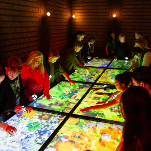 Personas reunidas alrededor de una mesa digital interactiva que muestra un mapa pixelado inspirado en un entorno de videojuego, en un espacio iluminado con tonos cálidos.