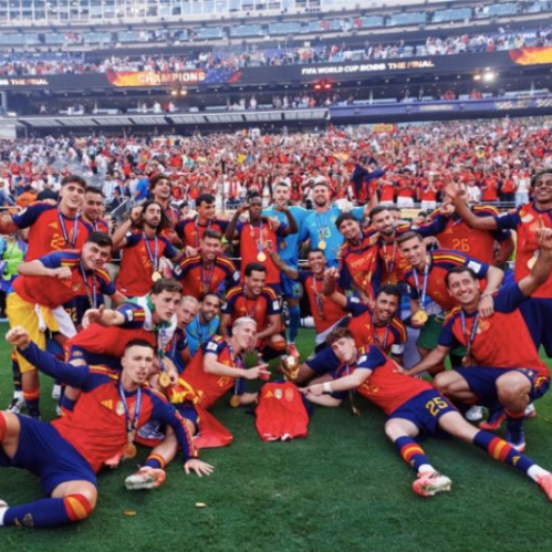 Fotografía deportiva en plano general en un estadio de fútbol iluminado con gradas llenas de aficionados. Al centro del campo de juego sobre el césped, los jugadores de la Selección Española celebran eufóricos vistiendo la camiseta roja oficial y medallas doradas al cuello; algunos posan acostados en el pasto, otros sentados y varios levantando las manos con expresiones de alegría junto a la réplica de la copa.