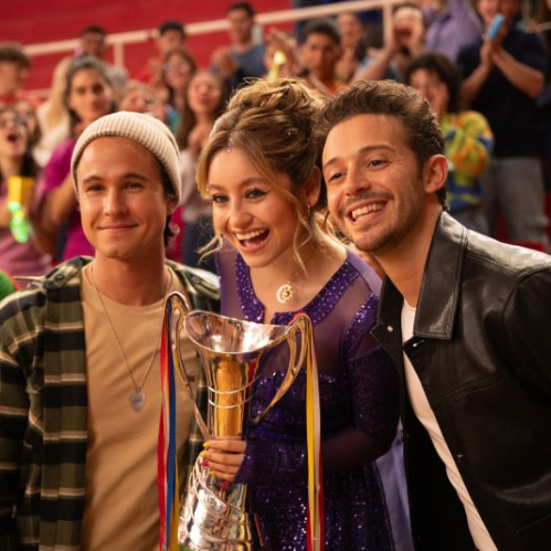 Fotografía de plano medio con iluminación cálida sobre un fondo de gradas rojas con público celebrando. Al centro, la actriz Karol Sevilla luce sonriente sosteniendo un trofeo de plata con cintas de colores; viste un vestido morado con pedrería y detalles brillantes. A su izquierda, Michael Ronda sonríe vistiendo una gorra tejida beige, playera beige y camisa de franela a cuadros; a su derecha, Ruggero Pasquarelli sonríe vistiendo una chaqueta de piel negra sobre playera blanca.