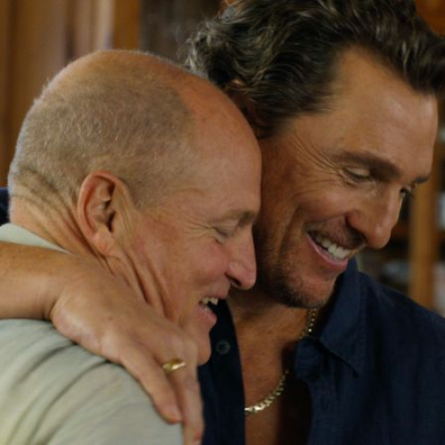 Fotograma de la serie de comedia 'Casi hermanos'. En primer plano, el actor Woody Harrelson, calvo y sonriendo ligeramente, abraza efusivamente al actor Matthew McConaughey, quien luce cabello ondulado castaño, sonríe cálidamente y viste una camisa de botones color azul oscuro con una cadena dorada al cuello. La escena se desarrolla en un interior iluminado de un rancho con estanterías de madera de fondo.