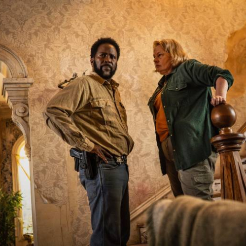 Dos personajes dentro de una casa deteriorada, en un ambiente tenso y oscuro que sugiere un escenario de misterio y peligro.