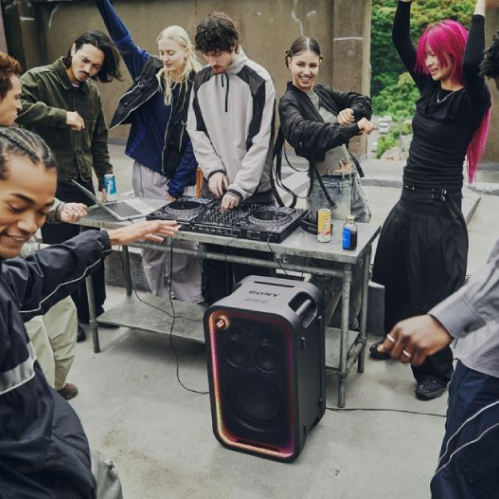 Un grupo de jóvenes baila y festeja alrededor de una mesa de DJ al aire libre mientras una bocina vertical Sony ULT TOWER reproduce música con iluminación multicolor en sus bordes.