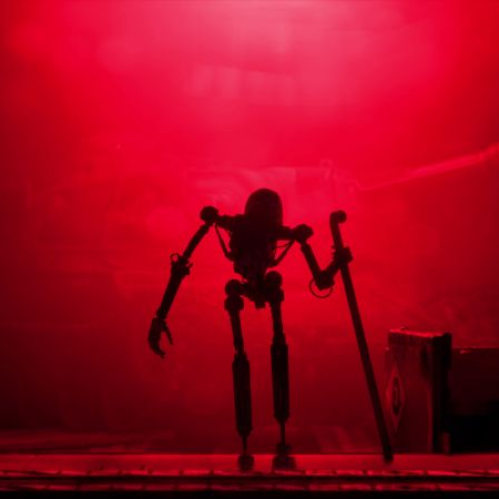 Fotografía en plano general con encuadre de marco que simula una puerta o escotilla, dominada por una intensa iluminación ambiental en color rojo saturado y neblina. En el centro, en silueta oscura y detallada, se observa una figura robótica o mecha humanoide estilizada, delgada y con extremidades articuladas, apoyada sobre un bastón largo metálico con su mano izquierda. Al fondo, a través de la densa atmósfera roja, se alcanza a distinguir la imponente figura de un tanque de guerra.