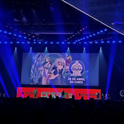 Escenario iluminado en tonos azules y morados con una pantalla gigante que muestra arte promocional de That Time I Got Reincarnated as a Slime: The Movie – Scarlet Bond, acompañado de banners laterales y público asistente.