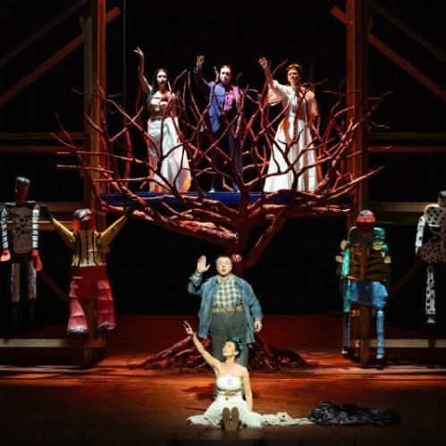 Escena teatral con un árbol escultórico rojo en el centro, rodeado de intérpretes y figuras humanas estilizadas en un escenario iluminado.