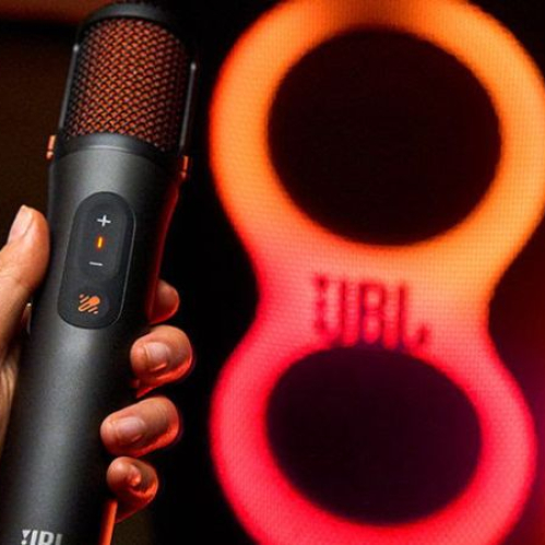 Un usuario sostiene con la mano un micrófono inalámbrico de color negro con la interfaz de controles iluminada en tono naranja. Al fondo se observa el panel de iluminación LED en forma de número ocho de un altavoz JBL PartyBox encendido en tonos rojos y naranjas.