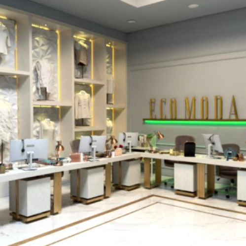 Fotografía en plano general que muestra las remodeladas oficinas de Ecomoda. En el fondo, sobre una pared gris, se lee el icónico logotipo "ECO MODA" en letras doradas iluminadas por una luz neón verde inferior. A la izquierda, un exhibidor de pared con nichos iluminados muestra varios maniquíes vistiendo sacos y prendas de alta costura en tonos blancos y crema. Al centro se extiende un largo escritorio blanco de trabajo modular con varias computadoras de escritorio, lámparas de tono cobre y sillas de oficina en color gris. A la derecha se aprecia un marco de puerta verde que da acceso a una sala contigua.