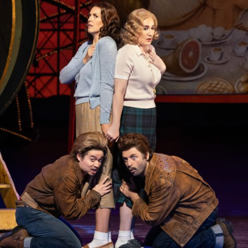 Fotografía de escena de la ópera Così fan tutte del Metropolitan Opera. En el escenario, dos actrices vestidas con vestuario retro de los años 50 se encuentran de pie espalda con espalda, mientras dos actores arrodillados a sus pies sostienen sus piernas con gesto dramático y cómico frente a un fondo decorado de feria.