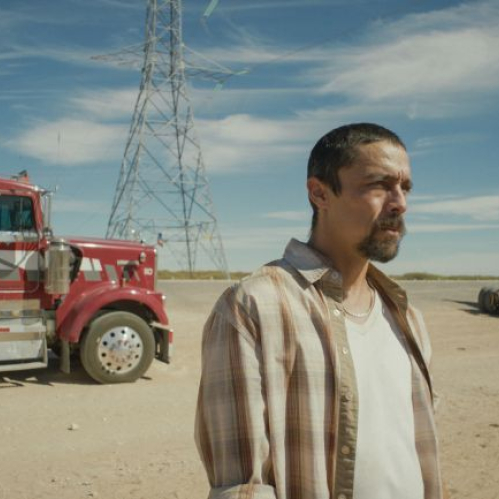 Fotograma en plano medio de la película "En el camino". En primer plano a la derecha aparece el actor Víctor Prieto luciendo bigote, barba de candado y vestimenta casual en tonos claros. De fondo, sobre un terreno árido bajo un cielo despejado, se observan un gran tráiler color rojo estacionado a la izquierda, la estructura de una torre de alta tensión al centro y un chasis de camión a la derecha.