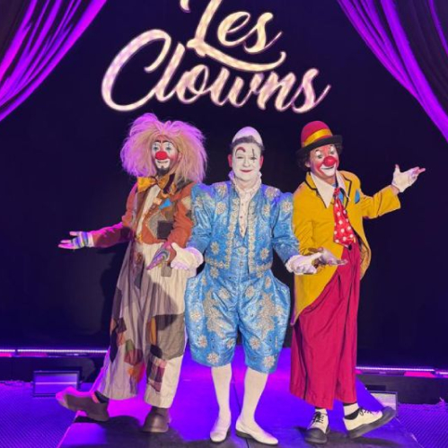 Tres artistas de clown caracterizados con vestuarios coloridos y maquillaje posan sobre el escenario frente a un fondo negro con el nombre del espectáculo 'Les Clowns' iluminado al centro y cortinas púrpuras a los costados. El personaje de la izquierda viste traje parcheado de tonos marrón y peluca rubia despeinada; el del centro luce un elegante traje azul claro de época con bordados dorados y rostro maquillado de blanco; y el de la derecha viste saco amarillo, pantalón rojo y sombrero rojo.