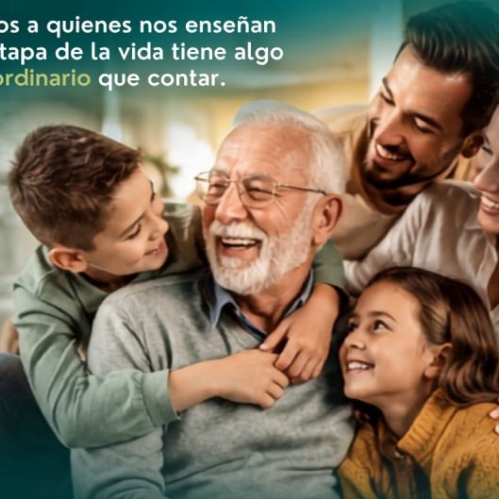 Fotografía cálida y familiar que muestra a un abuelo sonriente de cabello canoso y anteojos en el centro, abrazado efusivamente por sus nietos e hijos en el tono de una celebración familiar. En la esquina superior izquierda se lee el texto en letras blancas y verdes: 'Celebremos a quienes nos enseñan que cada etapa de la vida tiene algo extraordinario que contar.'