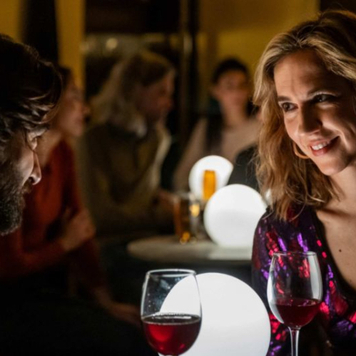 Dos personas conversan en un ambiente íntimo con iluminación cálida, acompañadas de copas de vino en una mesa.