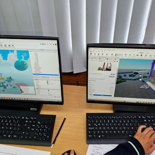 Dos monitores de computadora sobre un escritorio de madera mostrando la interfaz del software de desarrollo Roblox Studio durante un taller educativo, con un estudiante sosteniendo un lápiz y usando el teclado para diseñar mapas y niveles 3D interactivos.