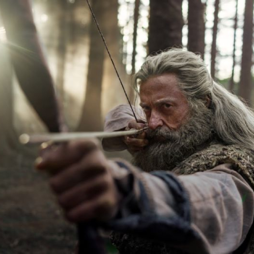 Fotograma de primer plano medio de Hugh Jackman caracterizado como Robin Hood en un bosque iluminado por destellos de luz solar entre los árboles. El actor luce cabello largo canoso, barba tupida con canas y ropas de época en tonos beige y pardo con textura gruesa. Jackman sostiene un arco de madera apuntando directo hacia la cámara con expresión concentrada y severa.
