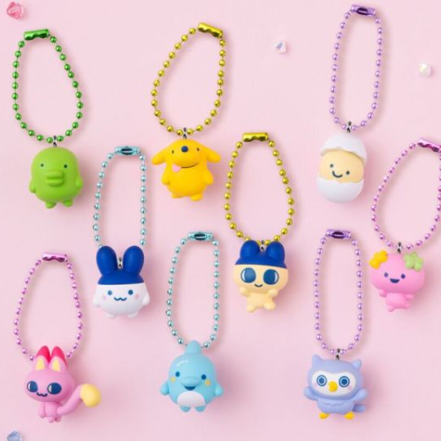 Colección de nueve llaveros de figuras estilo kawaii sobre un fondo rosa claro salpicado de pequeños cristales brillantes. Cada llavero presenta una criatura o personaje animado de colores vistosos como amarillo, azul, rosa, verde y morado, sujetados por una cadena de bolitas metálicas de colores combinados. En la esquina inferior derecha aparece el sello redondo oficial de Bandai Shokugan.