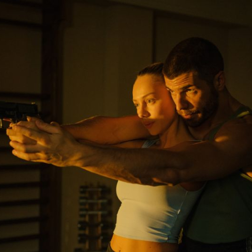 Fotografía en plano medio corto con iluminación cálida y lateral en un gimnasio. Muestra a Hugo Diego García abrazando por la espalda a Ester Expósito mientras sostiene sus manos para guiarla y enseñarle a apuntar con una pistola negra hacia el frente.