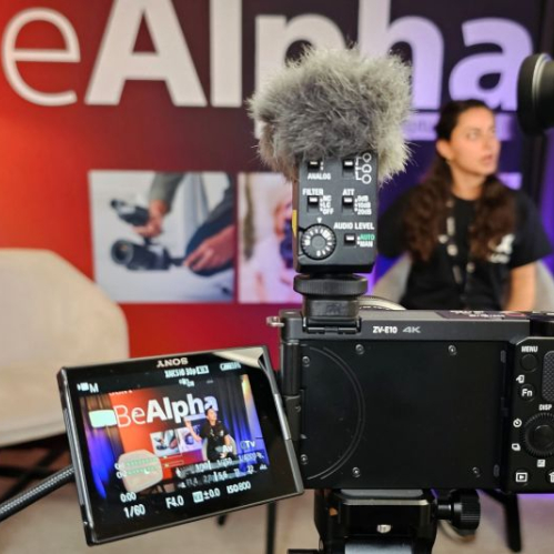 Cámara Sony ZV-E10 montada sobre un trípode con micrófono de cañón y pantalla abatible encendida con el logotipo Be Alpha, enfocando a una persona en un set de grabación.