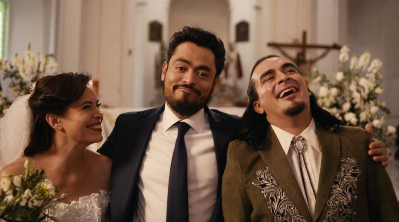 Novia vestida de blanco sonriendo junto a dos hombres en trajes elegantes, uno de ellos con una sonrisa exagerada, durante una ceremonia en el interior de una iglesia.