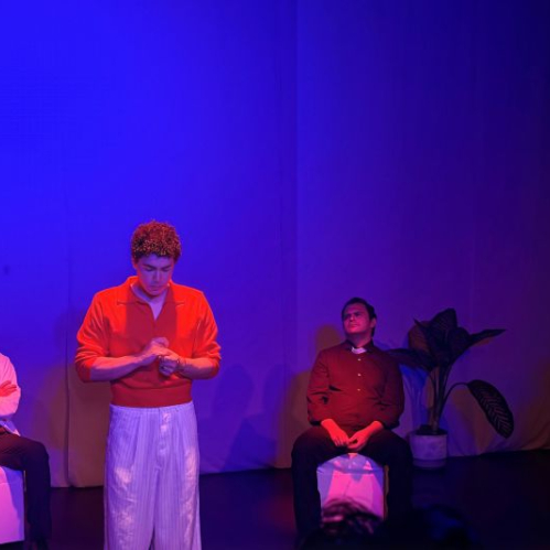 Elenco de la obra Di.OS en el escenario con iluminación en tonos azules y púrpuras, sentados y de pie junto a plantas en macetas en un entorno minimalista.