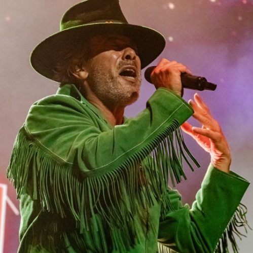 Jay Kay interpretando con pasión al micrófono vistiendo su chaqueta verde con flecos y sombrero oscuro sobre un fondo iluminado.