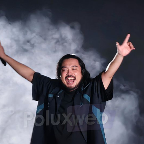Artista en escenario con los brazos levantados entre humo y luces dramáticas.