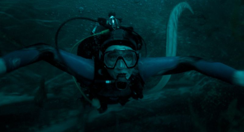 Austin Abrams en una escena de supervivencia bajo el agua en "La caída de la ballena". Fotografía en formato horizontal ambientada bajo el agua en tonos azul oscuro y verdoso. En el centro, el actor Austin Abrams aparece caracterizado como buzo, vistiendo un traje de neopreno con visor y boquilla de oxígeno mientras extiende los brazos nadando en medio de aguas profundas.