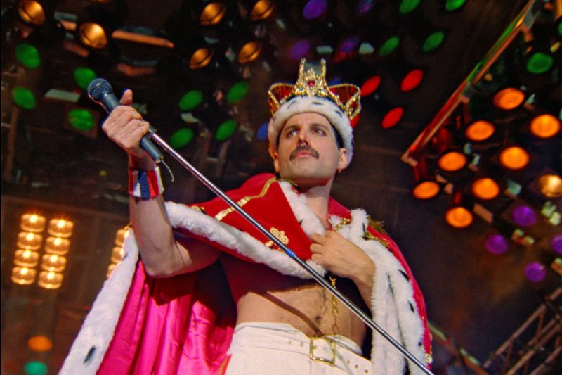 Freddie Mercury en una escena del concierto "Queen Budapest", restaurado en 4K. Fotografía en formato horizontal de una presentación en vivo de Queen. Al centro, el vocalista Freddie Mercury aparece de perfil usando una corona dorada con detalles rojos y armiño sobre la cabeza, luciendo una capa roja de felpa con bordes blancos sobre el torso descubierto y pantalones blancos. Sostiene el pedestal de su micrófono inclinado hacia abajo mientras las luces del escenario en tonos amarillos, verdes y rojos iluminan el fondo.
